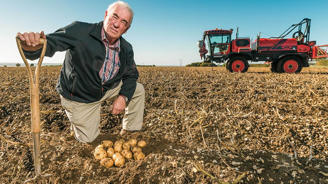 Robert kendal kneeling on farm next potatoes with a sprayer in the background